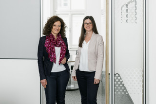 Zwei Damen aus dem Bereich HR stehen in einer offenen Glastür, blicken zur Kamera und lächeln.