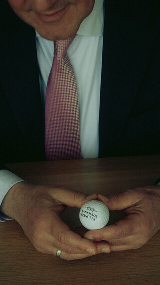 Nahaufnahme einer Person im dunklen Anzug, die einen Golfball mit dem Aufdruck „Bankhaus Spängler“ und Logo in den Händen hält.