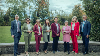 Acht Personen in Anzügen verschiedener Farben stehen draußen, mit einer Wiese und Bäumen im Hintergrund.