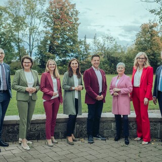 Acht Personen in Anzügen verschiedener Farben stehen draußen, mit einer Wiese und Bäumen im Hintergrund.