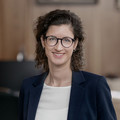 Frau mit braunem, lockigen Haar steht in einem Büro. Sie lächelt höflich und trägt einen dunklen Blazer mit einer weißen Spitzenbluse.