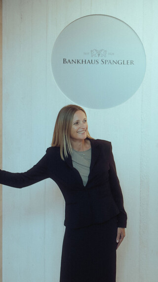 Person in Business-Kleidung steht neben einer Wand mit dem Bankhaus-Spängler-Logo und blickt nach rechts.