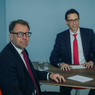 Dr. Nils Kottke, unscharf dargestellt, sitzt mit einem Mann an einem Tisch. Beide Männer in Anzügen mit roten Krawatten haben Unterlagen vor sich am Tisch.