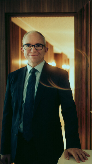 Ein Mann in Anzug, Krawatte und Brille blickt freundlich in die Kamera. Seine Hand ruht auf einem Sideboard.