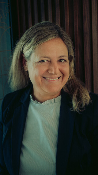 Eine blonde Frau lächelt. Sie trägt eine weiße Bluse mit Rüschen und einen dunklen Blazer.