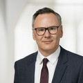 Der Oberkörper eines lächelnden Herren in Geschäftskleidung vor hellem Hintergrund.