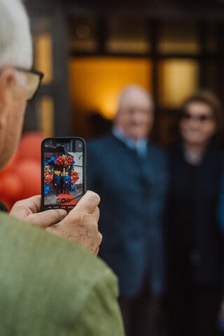 Blick über die Schulter eines älteren Herren, der mit dem Smartphone drei festlich gekleidete Personen fotografiert.