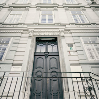 Fassade der Niederlassung Graz des Bankhaus Spängler. Graues Gebäude mit 4 Stockwerken, mehreren Fenster und einer schwarzen Tür. Ein Treppe mit Geländer dienen als Aufgang.