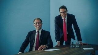 Zwei lachende Herren in Anzug an einem Tisch.