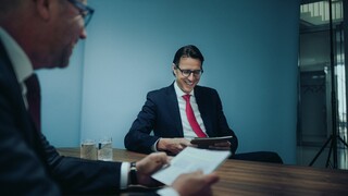 Dr. Nils Kottke sitzt einem unscharf dargestellten Mann gegenüber. Beide lächeln und halten Unterlagen in der Hand.