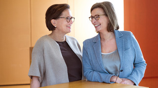 Zwei Damen stehen nebeneinander an einem Holzpult und lachen einander freundlich an. Beide haben kurze, braune Haare und tragen eine Brille. Eine Frau ist mit einem hellblauen Blazer gekleidet, die andere trägt einen beigefarbenen Blazer.