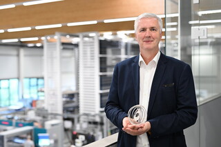 Ein Mann im Anzug blickt in die Kamera und hält ein kugelförmiges Objekt in der Hand, während im Hintergrund das Innere einer Fabrik zu sehen ist.