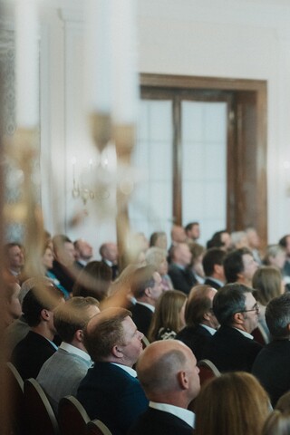 Elegant gekleidetes Publikum sitzt in einem historischen Saal unter einem Kronleuchter.