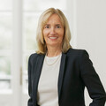 Frau mit blonden schulterlangen Haaren lächelt freundlich. Sie trägt eine weißes Bluse und einen schwarzen Blazer.