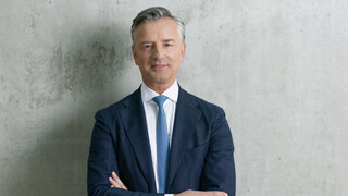 Mann mit grau-melierten Haaren und freundlichem Ausdruck steht vor einer grauen Wand. Er trägt ein weißes Hemd, eine hellblaue Krawatte und ein dunkelblaues Sakko. Er hat die Hände verschränkt.