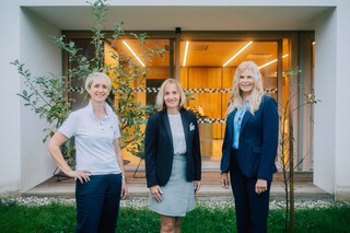 Drei Frauen lächeln in die Kamera, während sie draußen auf der Wiese vor einem Gebäude mit großen Fenstern und jungen Bäumen im Hintergrund stehen.