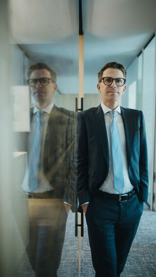 Person im Anzug mit Brille lehnt in einem modernen Büroflur an einer Wand. In einer Glaswand ist die Spiegelung der Person zu sehen.