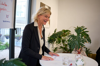 Eine Dame in Geschäftskleidung steht lachend an einem Stehtisch.