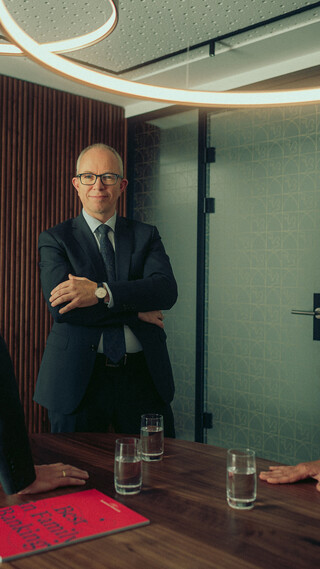 Drei Personen sitzen im Meetingraum und blicken einander an; auf dem Tisch liegt eine rote Broschüre.