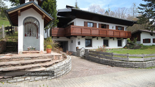 Zweistöckiges Einfamilienhaus von außen mit weißer Fassade, dunklem Holzbalkon und dunkelbraunen Fenstertafeln aus Holz. Eine Garage und eine kleine Hauskapelle sowie ein Garten sind erkennbar.
