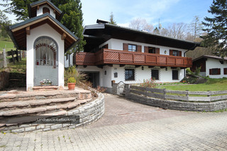 Zweistöckiges Einfamilienhaus von außen mit weißer Fassade, dunklem Holzbalkon und dunkelbraunen Fenstertafeln aus Holz. Eine Garage und eine kleine Hauskapelle sowie ein Garten sind erkennbar.
