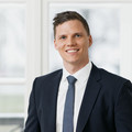Mann, stehend vor Fenster, lächelnd, kurze brünette Haare, in dunkelblauem Anzug, weißem Hemd und blauer Krawatte, die Hände in den Hostentaschen