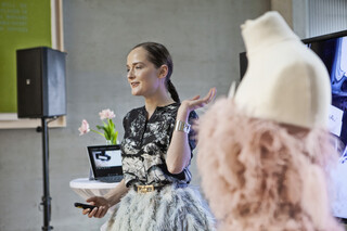 Eine exquisit gekleidete Dame hält neben einem ausgestellten Kleid einen Vortrag.