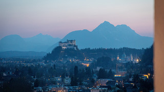 Panorama-Foto der Festung Hohensalzburg, im Hintergrund die Berge, es herrscht abendliche Stimmung, einzelne Gebäude sind beleuchtet