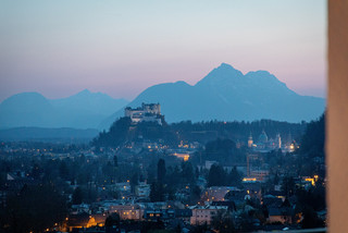 Panorama-Foto der Festung Hohensalzburg, im Hintergrund die Berge, es herrscht abendliche Stimmung, einzelne Gebäude sind beleuchtet