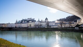 Blick von der Salzach unter einer geschwungenen Brücke auf die Salzburger Altstadt und die Festung Hohensalzburg im Hintergrund.