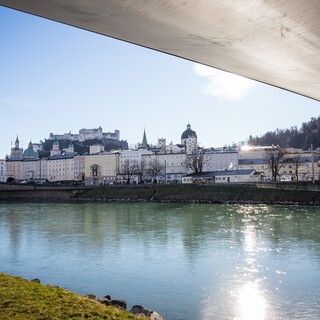 Blick von der Salzach unter einer geschwungenen Brücke auf die Salzburger Altstadt und die Festung Hohensalzburg im Hintergrund.