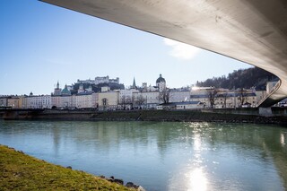 Blick von der Salzach unter einer geschwungenen Brücke auf die Salzburger Altstadt und die Festung Hohensalzburg im Hintergrund.
