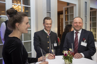 Drei Personen (2 Männer, 1 Frau) stehen bei einem Stehtisch mit weißer Husse und rotem Blumen-Arrangement. Sie halten ein Getränk mit Strohhalm und Limette in der Hand. Alle Personen lächeln. Die Frau trägt ein schwarzes Oberteil und hat hochgesteckte Haare. Ein Mann trägt einen Trachtenanzug, der andere einen Anzug mit weißem Hemd und dunkelrot-gestreifter Krawatte. 