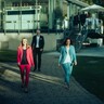 Zwei Damen und ein Herr in Geschäftskleidung gehen fröhlich auf einem gepflasterten Weg von einem Gebäude mit Wintergarten weg.