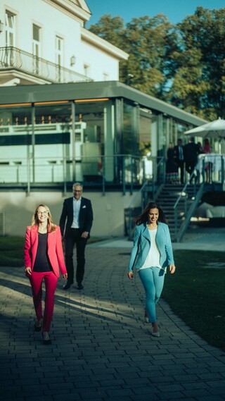 Zwei Damen und ein Herr in Geschäftskleidung gehen fröhlich auf einem gepflasterten Weg von einem Gebäude mit Wintergarten weg.