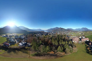 Panoramabild von oben mit grüner Wiese, herbstlich gefärbten Bäumen und mehreren Gebäude. Im Hintergrund sind Berge, ein blauer Himmel und die untergehende Sonne zu sehen.
