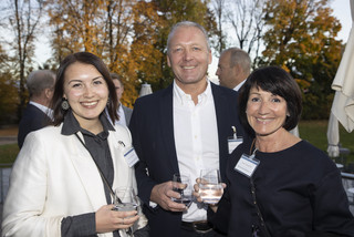 Zwei Frauen und ein Mann lächeln, alle haben ein Glas Wasser in der Hand und sind schwarz-weiß gekleidet. Sie befinden sich im Freien und im Hintergrund sind orange-leuchtende Bäume zu sehen.