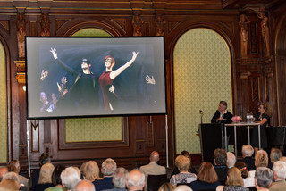 Menschen blicken auf eine Leinwand, auf der ein Bild mit zwei tanzenden Personen zu sehen ist