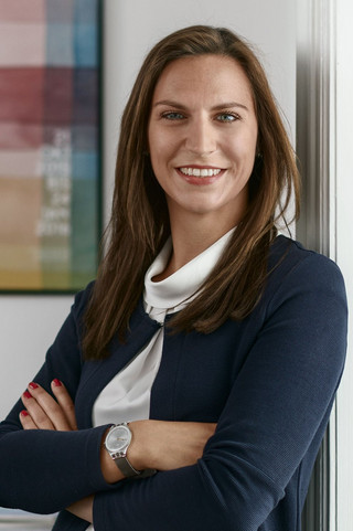 Ein Frau lehnt am Türrahmen in einem Bürogebäude des Bankhaus Spängler. Sie lächelt freundlich und hat lange, braune Haare. Im Hintergrund ist ein buntes Bild ersichtlich. 