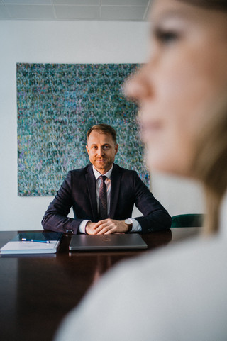 Am Tisch sitzt ein Mann im blauen Anzug, die Hände auf dem Tisch. 