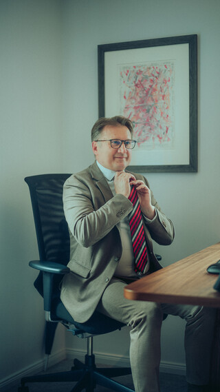 Ein Mann mit grauen Haaren und Brille sitzt in einem braunen Anzug mit rot gestreifter Krawatte auf einem Bürostuhl an seinem Schreibtisch und richtet seine Krawatte.
