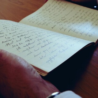Hände halten ein aufgeschlagenes Notizbuch mit handschriftlichen Notizen.