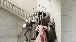 Webesujet des Bankhaus Spängler: Eine dunkel-gekleidete Familie mit Schwan-Köpfen steht auf einer Treppe. Eine Dame sticht besonders hervor - sie hat einen Flamingo-Kopf und trägt ein opulentes Kleid in rosa Farbe.