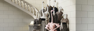 Webesujet des Bankhaus Spängler: Eine dunkel-gekleidete Familie mit Schwan-Köpfen steht auf einer Treppe. Eine Dame sticht besonders hervor - sie hat einen Flamingo-Kopf und trägt ein opulentes Kleid in rosa Farbe.