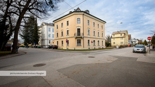 Historisches Gebäude mit 4 Stockwerken von außen, das sich an einer Straßenkreuzung befindet. Es hat eine dunkelgelbe Fassade und zahlreiche Fenster. Außerdem sind im Hintergrund weitere Häuser und ein paar parkende Autos zu sehen.