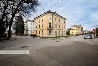 Historisches Gebäude mit 4 Stockwerken von außen, das sich an einer Straßenkreuzung befindet. Es hat eine dunkelgelbe Fassade und zahlreiche Fenster. Außerdem sind im Hintergrund weitere Häuser und ein paar parkende Autos zu sehen.