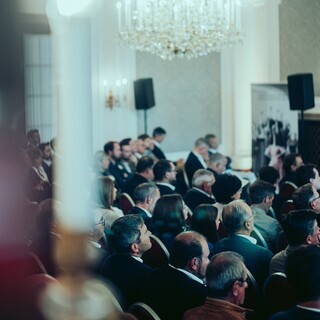 Publikum in Geschäftskleidung unter dem Kronleuchter eines historischen Saales.