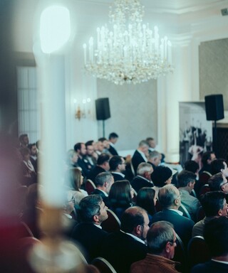 Publikum in Geschäftskleidung unter dem Kronleuchter eines historischen Saales.