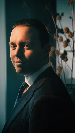 Ein dunkelhaariger Mann in dunklem Anzug und Krawatte blickt an der Kamera vorbei; im Hintergrund stehen Blumen.