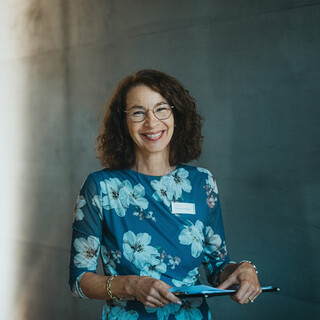 Eine Frau mit Blumenkleid und Brille blickt lächelnd in die Kamera und hält ein Klemmbrett in den Händen.